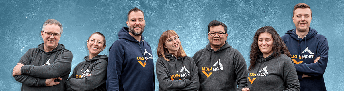 Ein Gruppenfoto von sieben lächelnden Mitarbeitern von LogiTel, die alle Sweatshirts mit dem Aufdruck "MOIN MOIN!" tragen und vor einem blauen Hintergrund stehen.