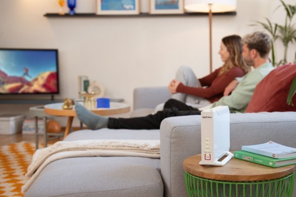 Ein weißer FRITZ!Box‑Router steht auf einem kleinen Beistelltisch in einem gemütlichen Wohnzimmer, während zwei Personen auf dem Sofa sitzen und eine Fernsehsendung ansehen.