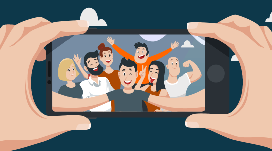 Im Vordergrund ist ein Smartphone zu sehen, das von zwei Händen gehalten wird. Auf dem Display ist eine Gruppe von Menschen zu sehen, die in die Handykamera blicken, um ein Gruppen-Selfie zu machen.