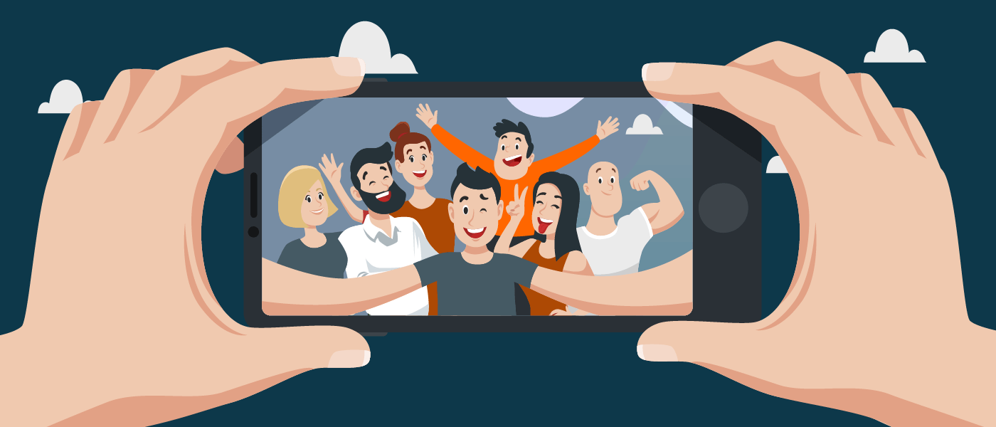 Im Vordergrund ist ein Smartphone zu sehen, das von zwei Händen gehalten wird. Auf dem Display ist eine Gruppe von Menschen zu sehen, die in die Handykamera blicken, um ein Gruppen-Selfie zu machen.
