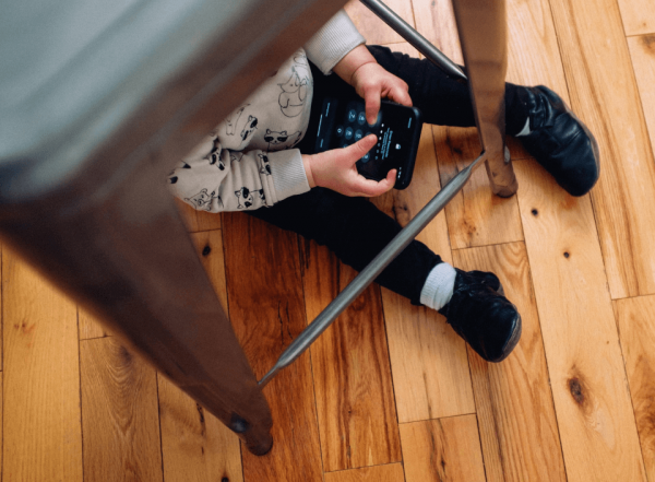 Ein Kleinkind sitzt unter einem Hocker und spielt mit einem Smartphone.