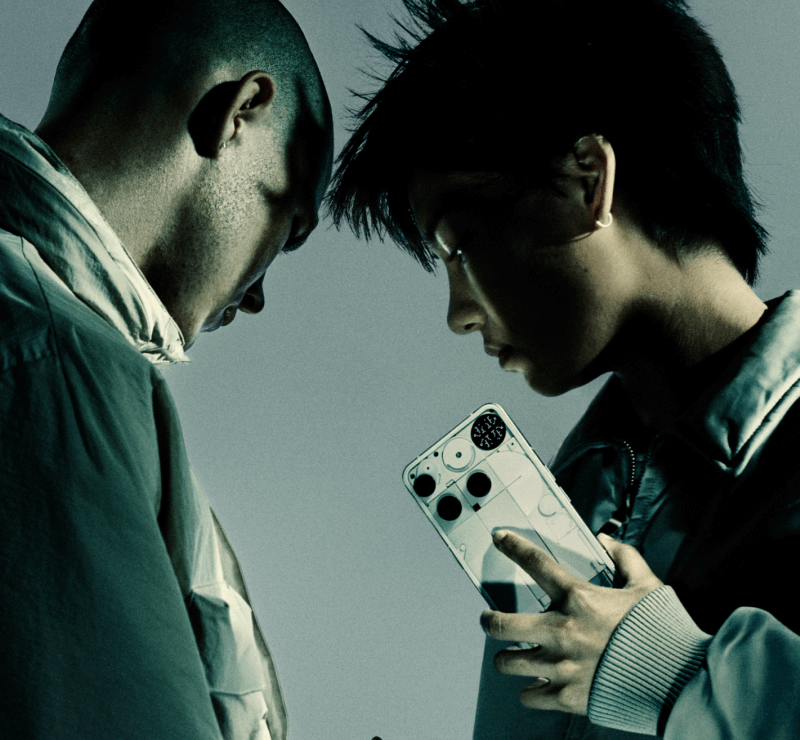 Zwei männliche Personen im Profil rechts und links am Bildrand. Die rechte Person hält in der linken Hand das Nothing Phone (3) in der Farbe weis, sodass Glyth-Rückseite zu sehen ist.