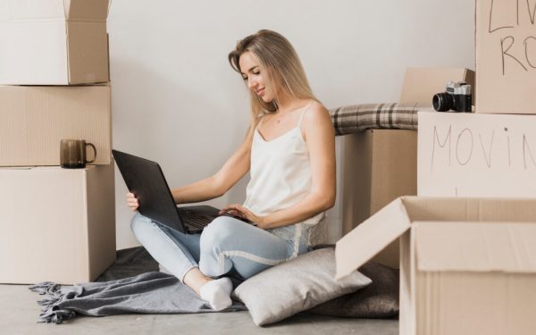 Eine junge Frau sitzt zwischen Umzugskartons am Laptop.