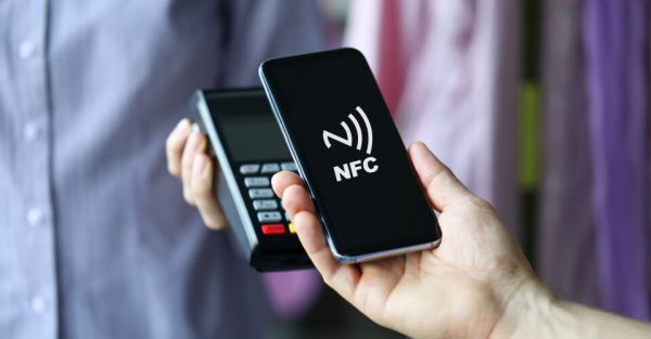 Ein Smartphone wird vor ein NFC-Lesegerät gehalten. Auf dem Display des Handys ist das Wellensymbol für die NFC-Technologie zu sehen. 