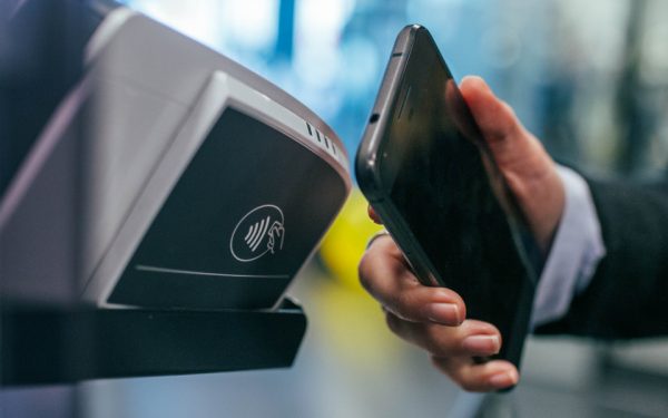 Ein Smartphone wird an ein NFC-fähiges Kassenterminal gehalten. Auf dem Lesegerät ist das Wellensymbol der Funk-Technologie zu sehen.