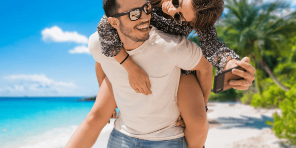 Lachende Frau ist huckepack auf dem Rücken eines Mannes am Strand, beide tragen Sonnenbrillen, im Hintergrund ist ein Palmenblatt. Zusätzlich ist der Text