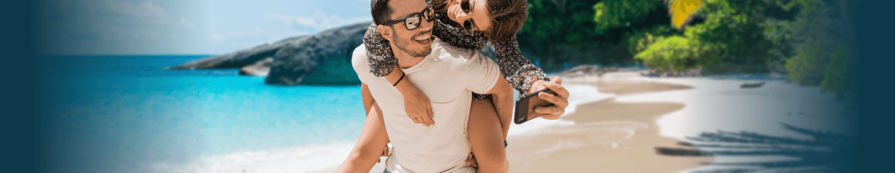 Lachende Frau ist huckepack auf dem R�cken eines Mannes am Strand, beide tragen Sonnenbrillen, im Hintergrund ist ein Palmenblatt. Zus�tzlich ist der Text 