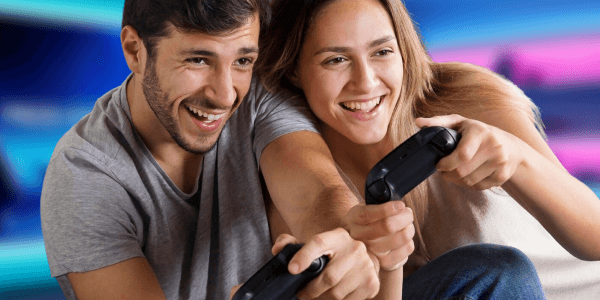 Zwei junge Menschen sitzen nebeneinander, lachen und spielen konzentriert mit Gamecontrollern ein Videospiel.