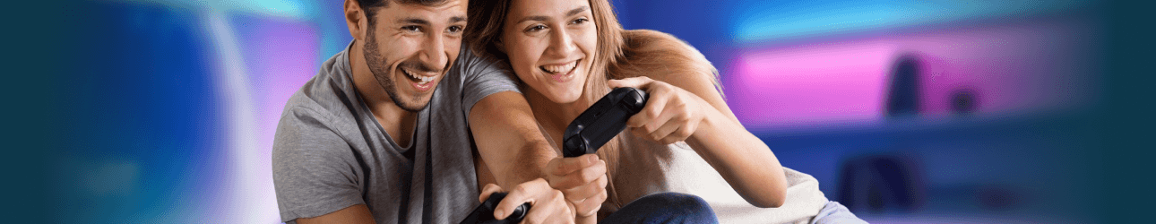 Zwei junge Menschen sitzen nebeneinander, lachen und spielen konzentriert mit Gamecontrollern ein Videospiel.