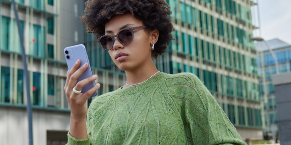 Junge Frau mit Sonnenbrille und grünem Strickpullover steht draußen vor einem modernen Bürogebäude und schaut konzentriert auf ihr iPhone..