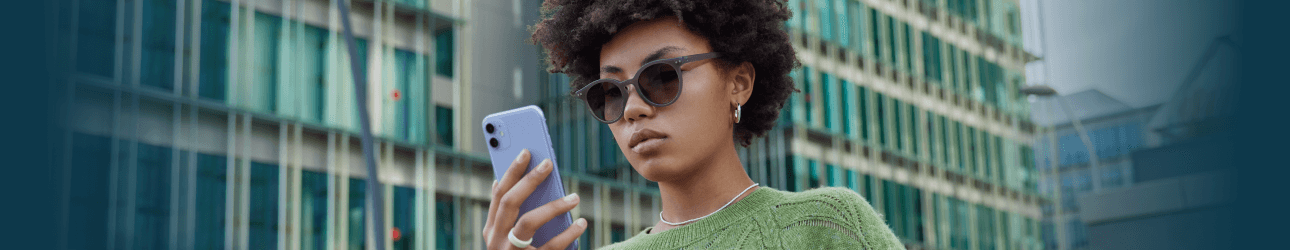 Junge Frau mit Sonnenbrille und gr�nem Strickpullover steht drau�en vor einem modernen B�rogeb�ude und schaut konzentriert auf ihr iPhone..