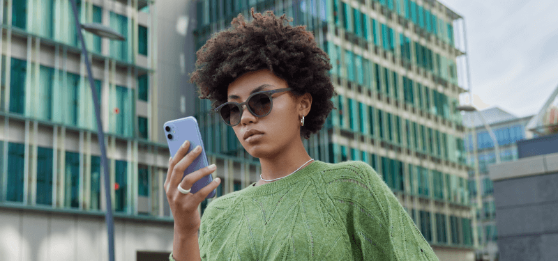 Junge Person mit Sonnenbrille und grünem Pullover blickt konzentriert auf ein Smartphone, im Hintergrund moderne Glasfassaden.
