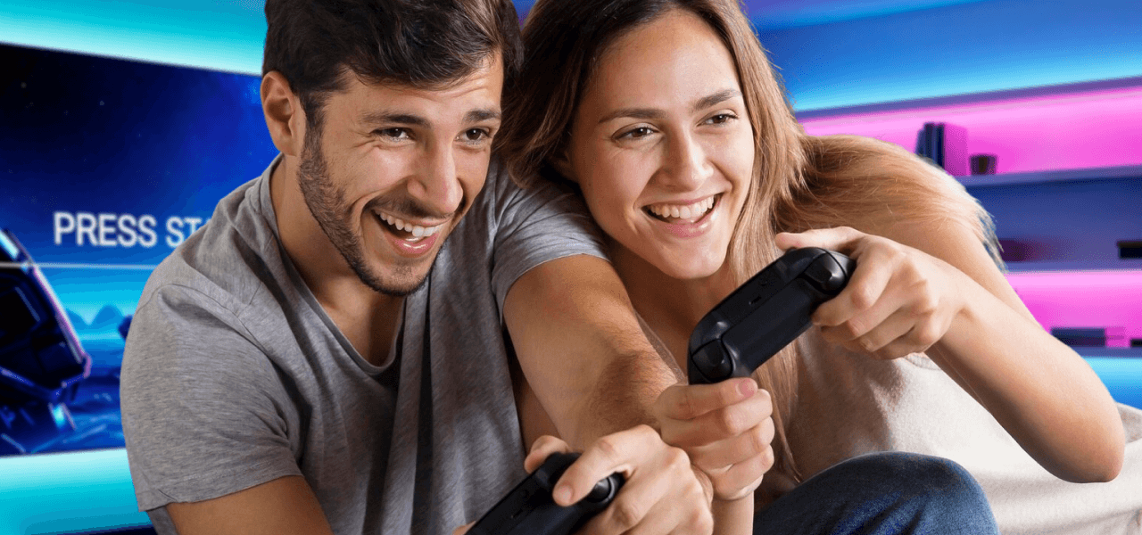 Zwei junge Menschen sitzen nebeneinander, lachen und spielen konzentriert mit Gamecontrollern ein Videospiel.