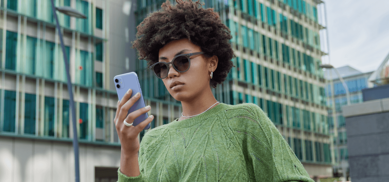 Junge Person mit Sonnenbrille und grünem Pullover blickt konzentriert auf ein Smartphone, im Hintergrund moderne Glasfassaden.