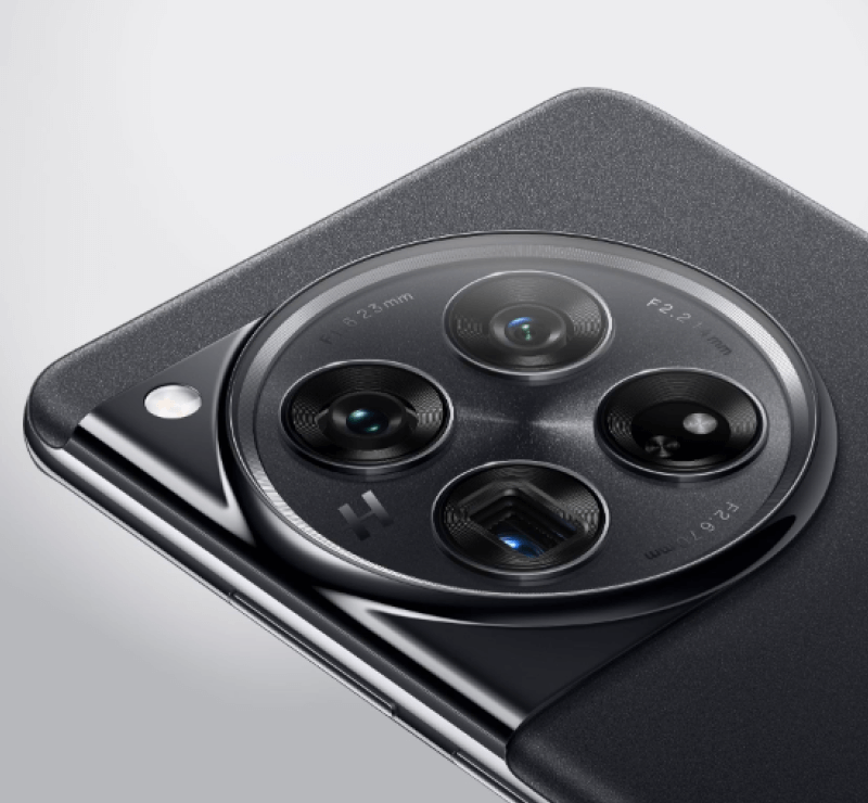 Nahaufnahme der Rückseite des OnePlus 12, mit Fokus auf das markante kreisförmige Kameramodul. Zu sehen sind vier Objektive, die symmetrisch angeordnet sind, sowie der Schriftzug "H", der auf die Zusammenarbeit mit Hasselblad hinweist. Das Kameramodul ist leicht erhaben und hebt sich glänzend vom matten, dunklen Gehäuse des Smartphones ab. Links daneben befindet sich ein LED-Blitz.